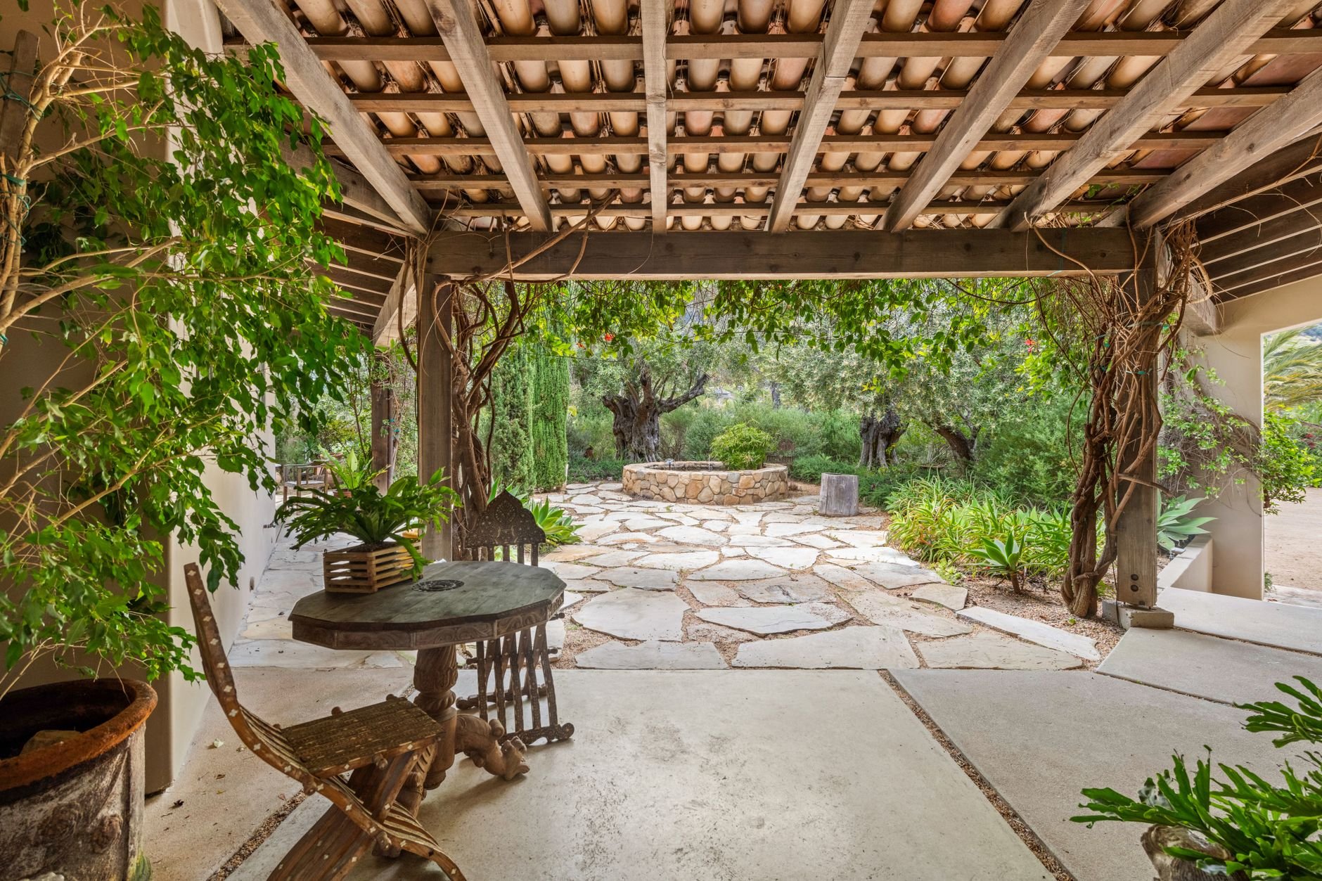 Rustic patio with wooden furniture, lush plants, and stone pathway in serene garden setting.